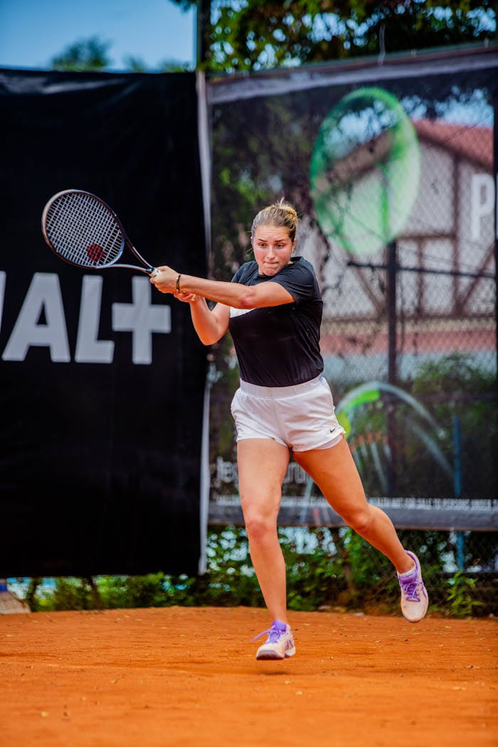 A female athlete playing tennis on a clay court with a powerful backhand stroke.